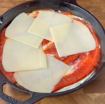 A cast iron skillet filled with a layer of tomato sauce and five slices of white cheese placed on top, sitting on a wooden surface.