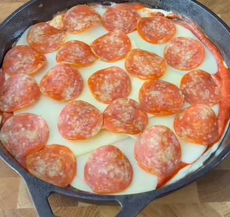 A cast iron skillet filled with a layer of pizza sauce, melted cheese slices, and pepperoni slices, resembling a deep dish or skillet pizza on a wooden surface.