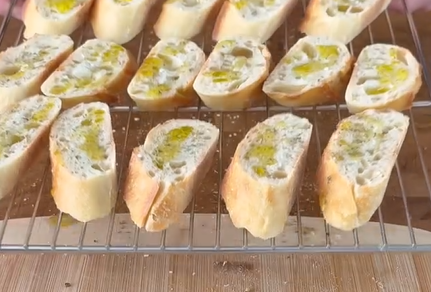 Slices of baguette topped with herbs and drizzled with olive oil are arranged on a wire rack, ready to be toasted. The rack is placed on a wooden surface.