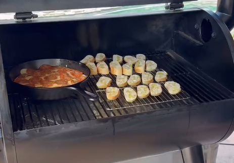 A black grill with a cast iron skillet containing meatballs in sauce on the left and sliced bread pieces topped with cheese arranged on the right side of the grill rack.