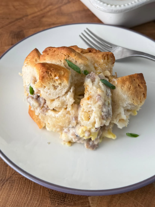A serving of biscuit casserole with sausage and eggs is on a white plate with a fork, resting on a wooden surface. The casserole is topped with chopped green onions.