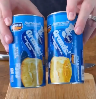 Two hands hold two blue cans of buttermilk biscuit dough, labeled Grands! with an image of a biscuit and the Pillsbury Doughboy on each can, above a wooden surface.