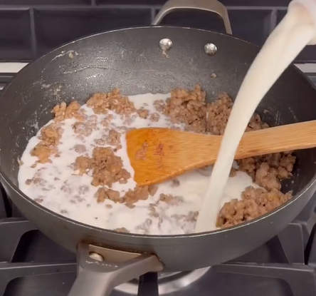 Ground sausage is being cooked in a skillet on a stove, while milk is being poured in and a wooden spatula is used to stir the mixture.