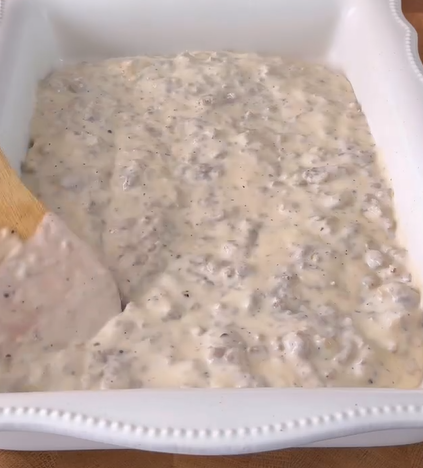 A wooden spoon spreads a creamy mixture with ground meat and black pepper in a white rectangular ceramic baking dish.