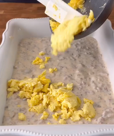 A spatula is transferring scrambled eggs from a pan into a white baking dish filled with a creamy sausage gravy mixture.