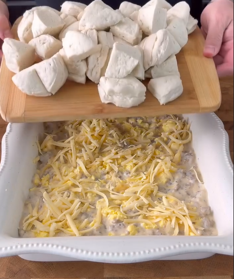 A person holds a wooden board with chunks of biscuit dough above a white baking dish filled with a creamy mixture topped with shredded cheese, preparing to add the dough pieces.
