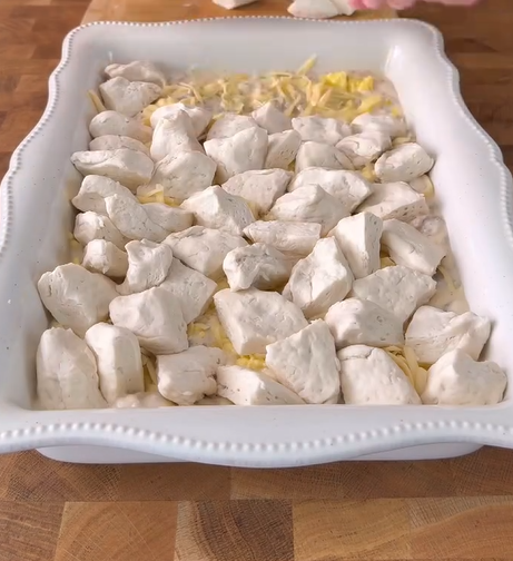A white rectangular baking dish on a wooden surface contains raw, evenly spaced biscuit dough pieces on top of shredded cheese.