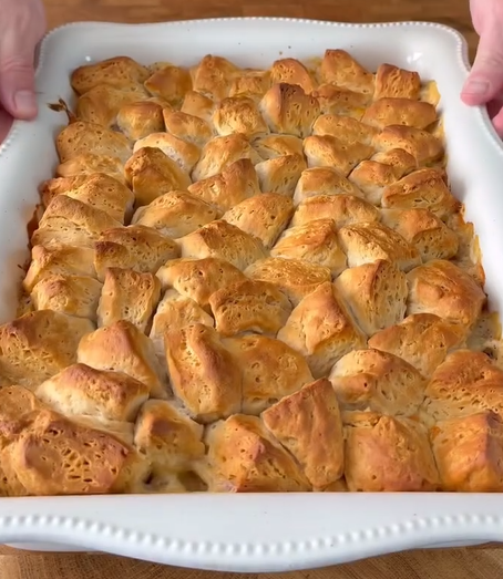 A white ceramic baking dish filled with golden-brown, baked biscuit pieces, cut into bite-sized chunks and arranged close together, sits on a wooden surface. Hands are holding the edges of the dish.