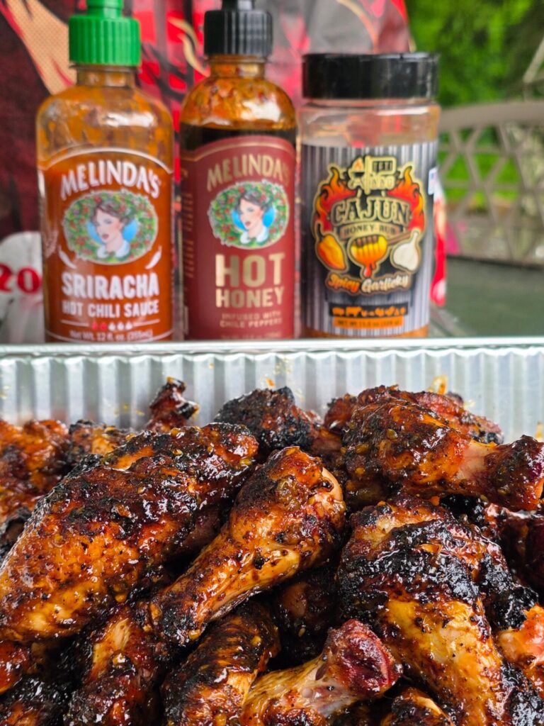 Grilled chicken drumsticks with a caramelized glaze in a foil tray, in front of bottles of Melinda’s Sriracha Hot Chili Sauce, Melinda’s Hot Honey, and a Cajun Spice Certified seasoning.