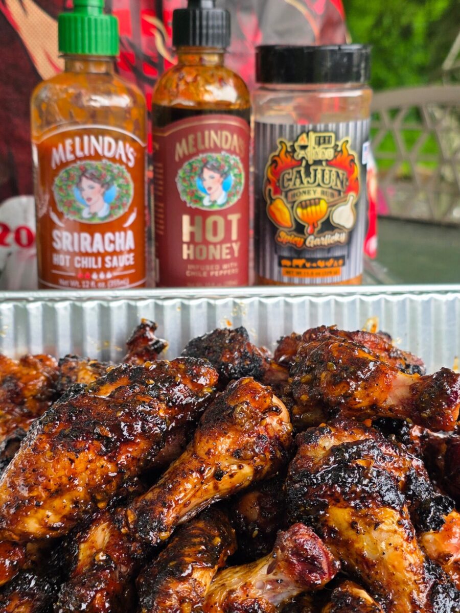 Grilled chicken drumsticks with a caramelized glaze in a foil tray, in front of bottles of Melinda’s Sriracha Hot Chili Sauce, Melinda’s Hot Honey, and a Cajun Spice Certified seasoning.