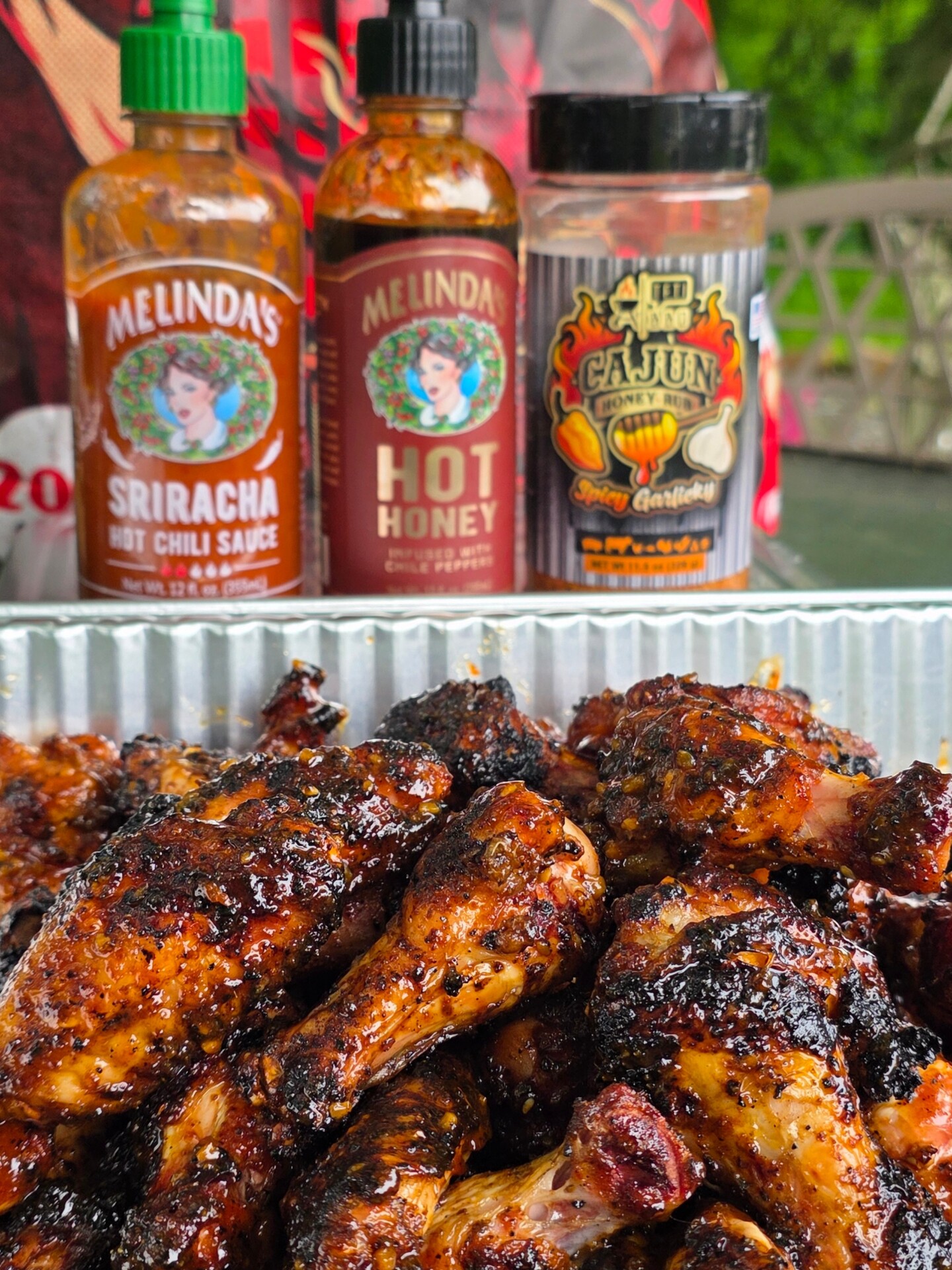 Grilled chicken drumsticks with a caramelized glaze in a foil tray, in front of bottles of Melinda’s Sriracha Hot Chili Sauce, Melinda’s Hot Honey, and a Cajun Spice Certified seasoning.