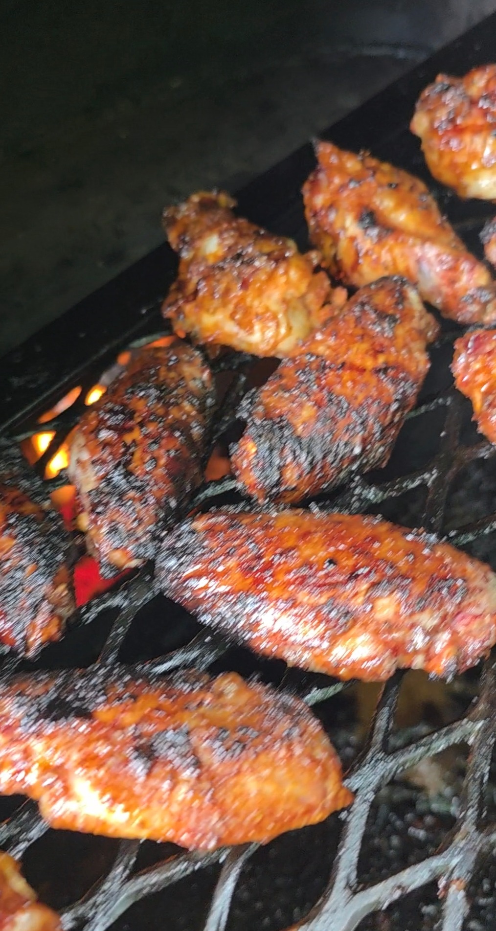 Several chicken wings are cooking on a grill over open flames, showing a charred, crispy exterior with a reddish-brown, smoky appearance. The grill grates are visible, with glowing embers beneath.