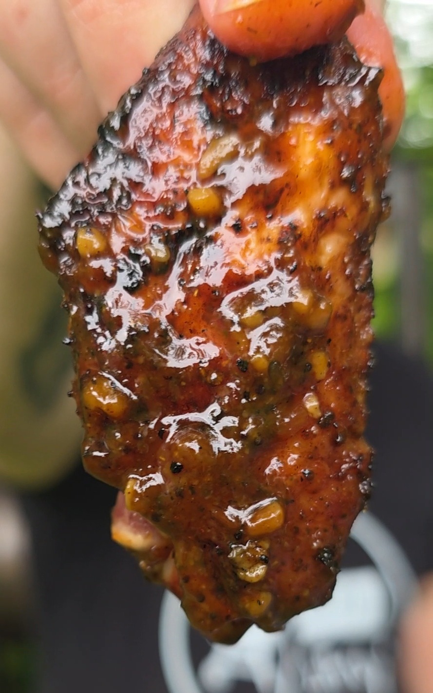 A close-up of a hand holding a glazed, grilled chicken wing with a shiny, spicy sauce coating and visible seasoning. The background is blurred.