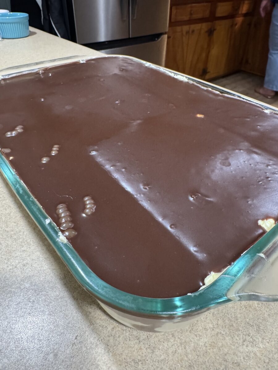 A glass baking dish filled with a dessert topped with a smooth layer of chocolate, sitting on a kitchen counter. A person stands in the background near wooden cabinets and a stainless steel refrigerator.