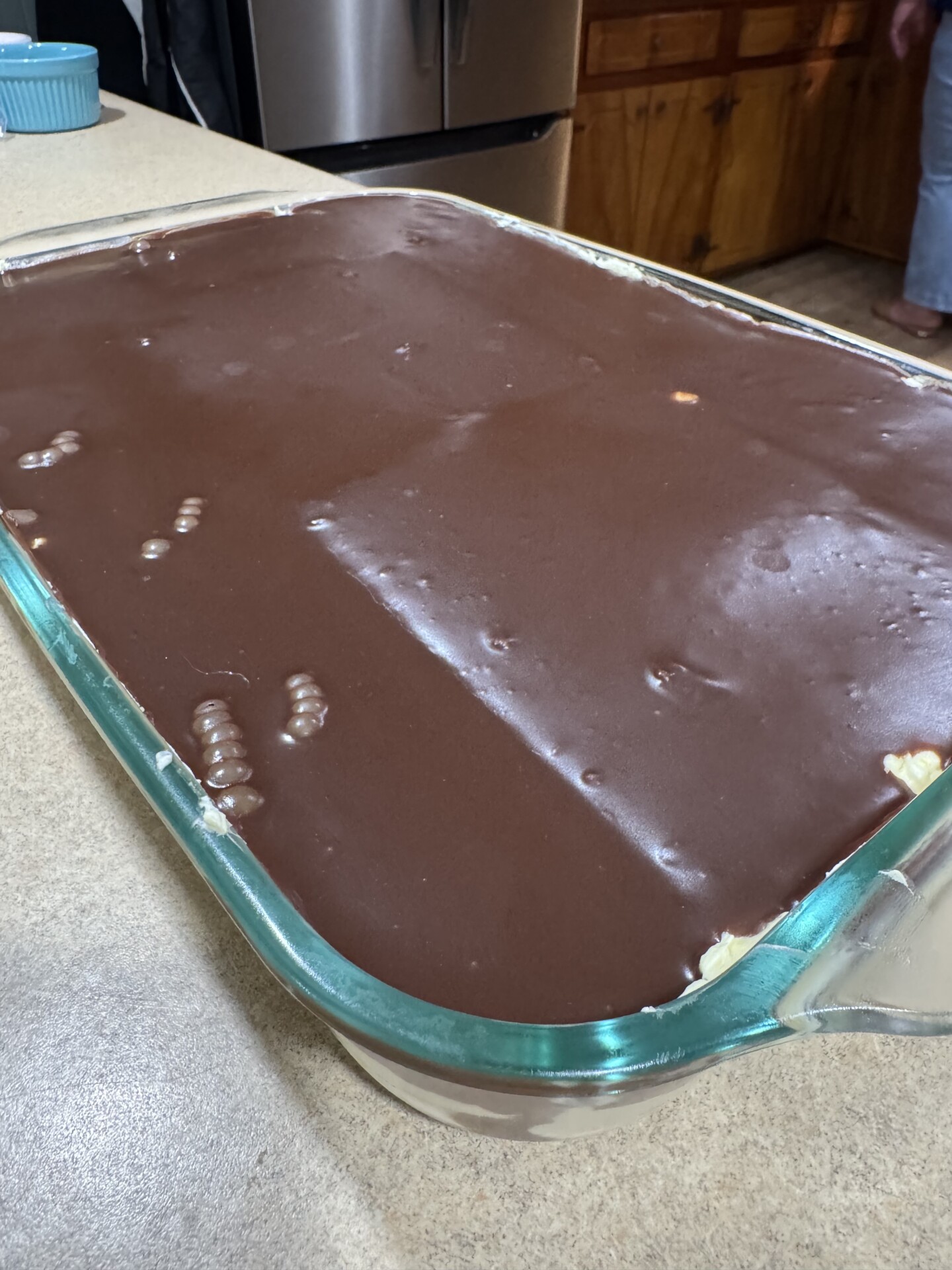 A glass baking dish filled with a dessert topped with a smooth layer of chocolate, sitting on a kitchen counter. A person stands in the background near wooden cabinets and a stainless steel refrigerator.