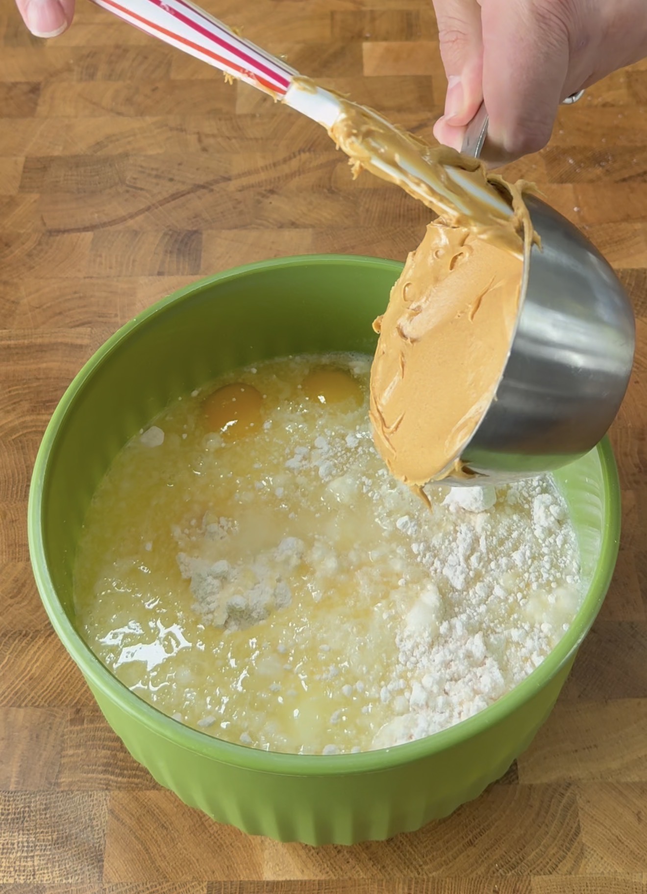 A hand is adding a cup of peanut butter to a green mixing bowl containing eggs, melted butter, and flour on a wooden countertop.