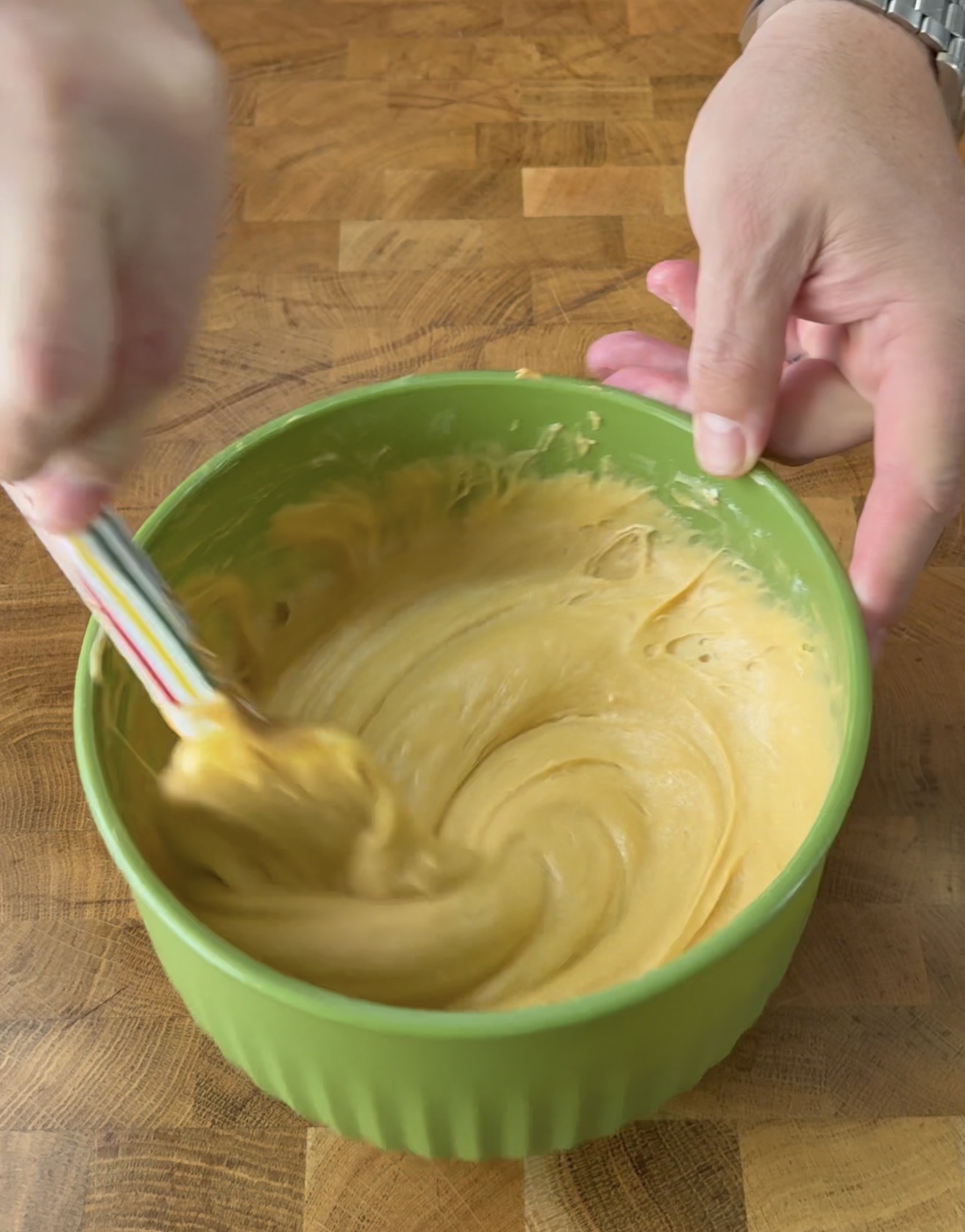 A person stirs thick, pale yellow batter in a green mixing bowl with a striped spatula on a wooden countertop.