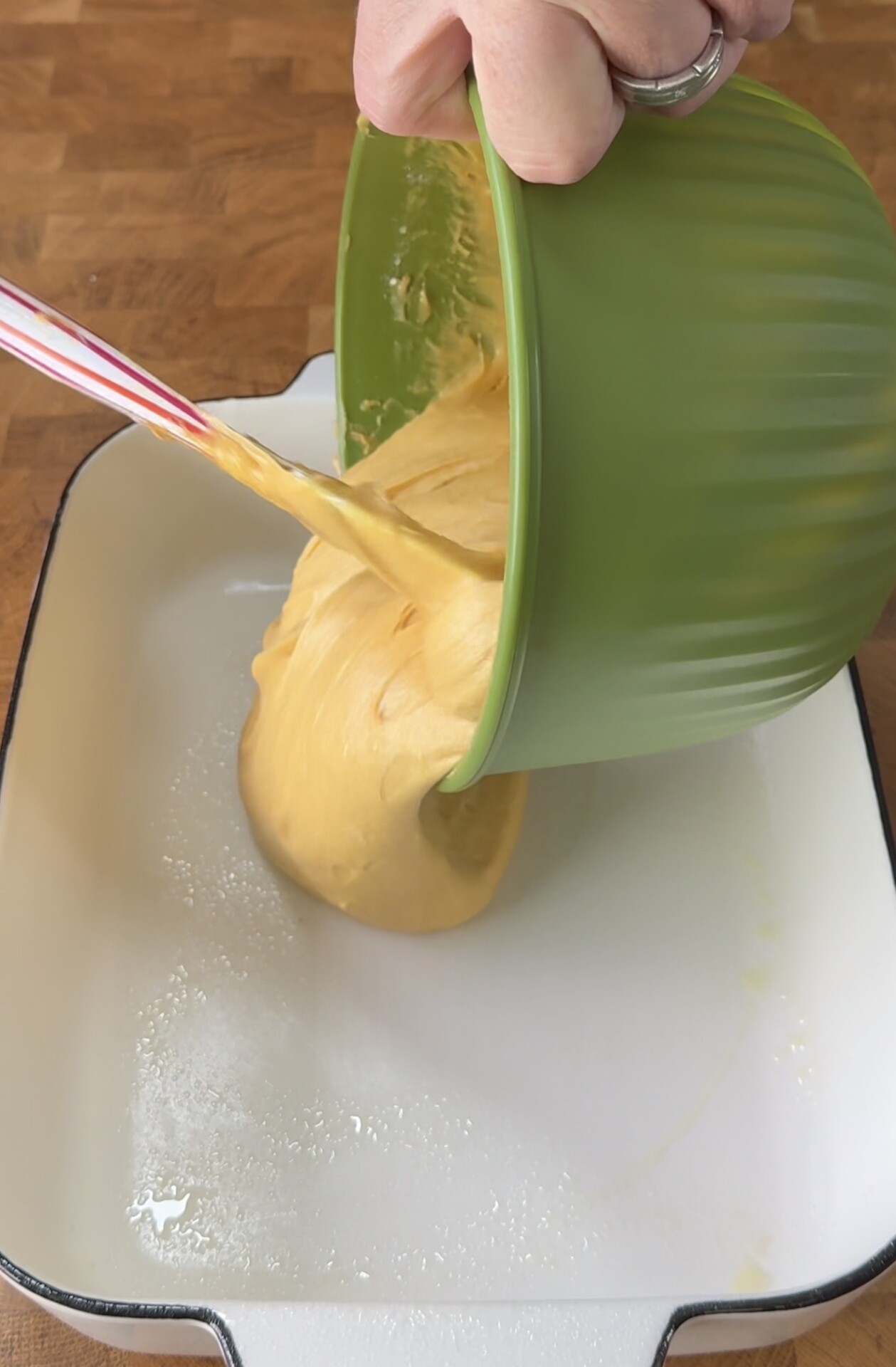 A hand holds a green bowl, pouring thick yellow batter into a greased white baking dish on a wooden surface. A red and white spatula helps guide the batter out of the bowl.