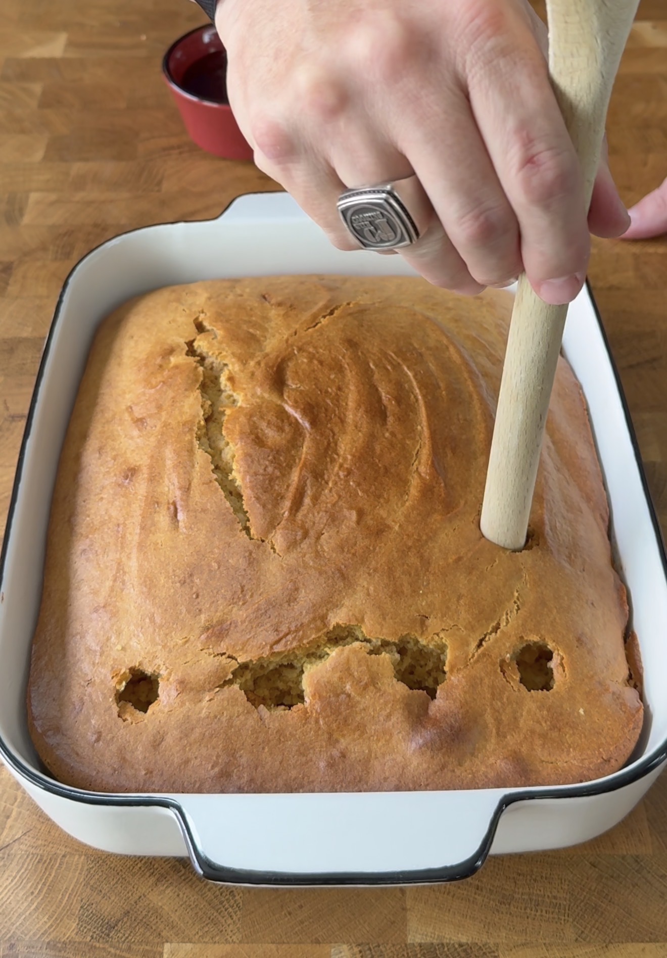 A hand holding a wooden spoon is poking holes into a freshly baked rectangular cake in a white baking dish on a wooden surface.