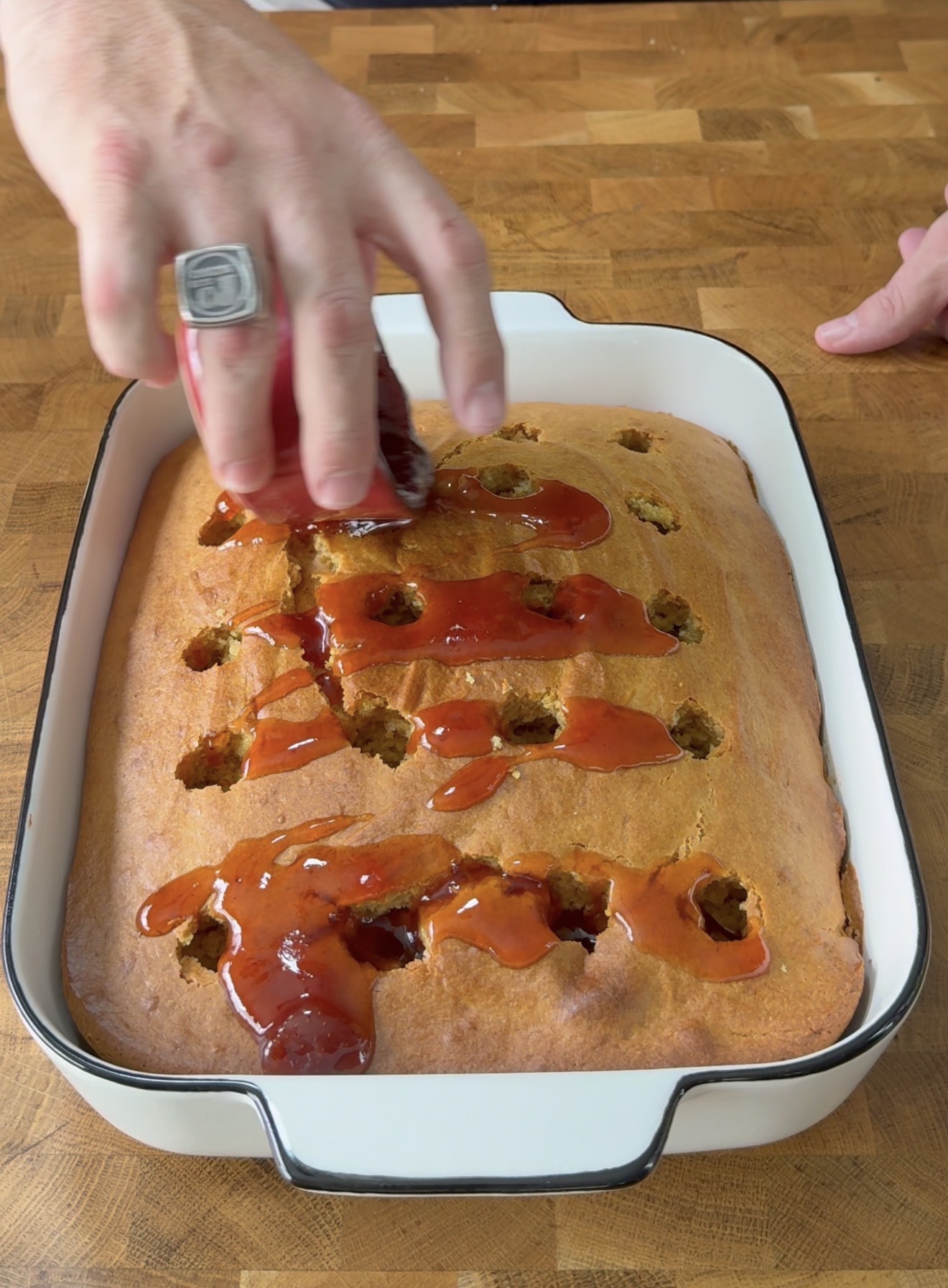 A hand spreads red sauce over a rectangular golden cake with holes in it, in a white baking dish on a wooden countertop.