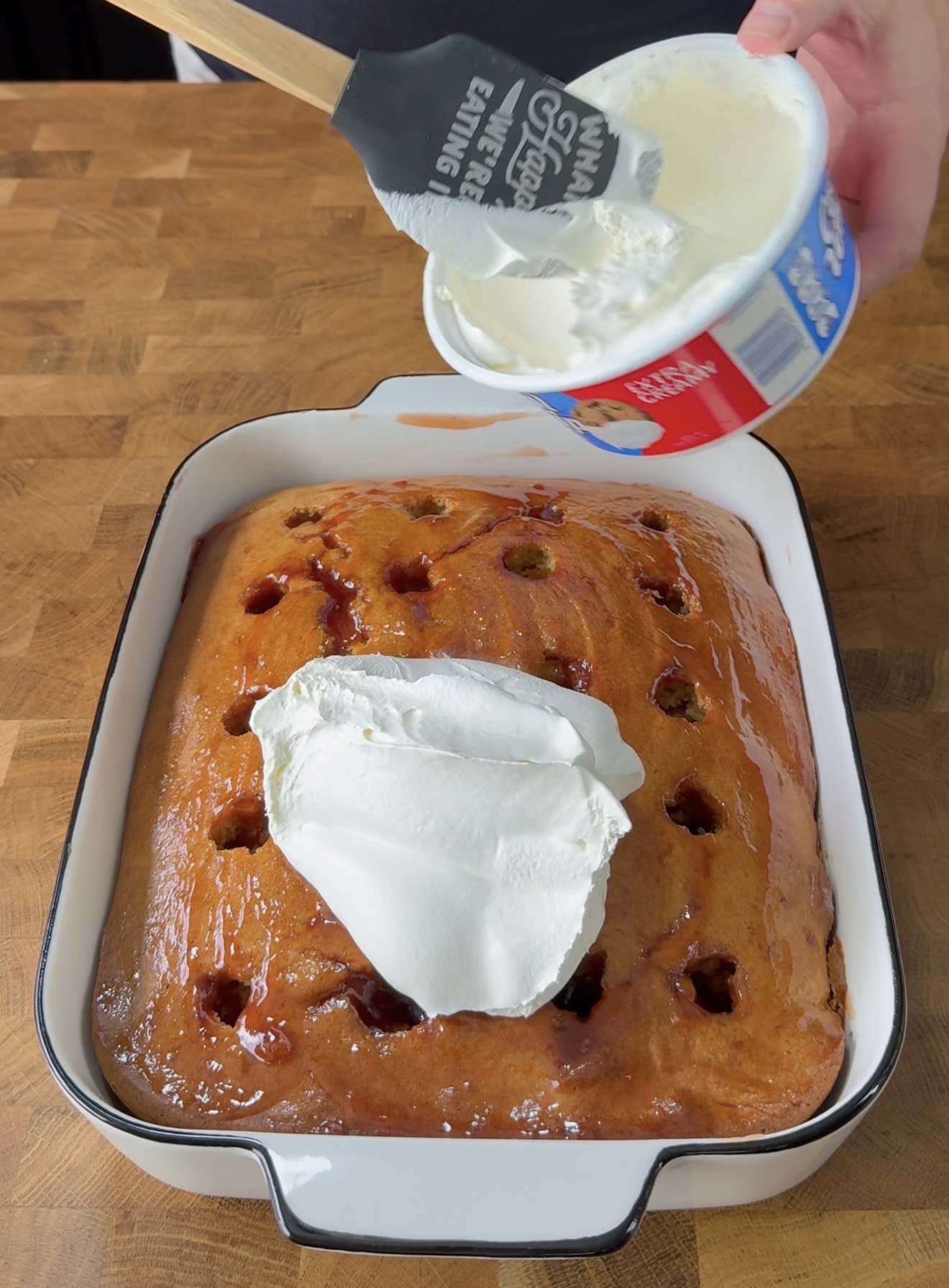 A hand is spreading whipped topping from a tub onto a rectangular cake with holes, set in a white baking dish on a wooden surface.
