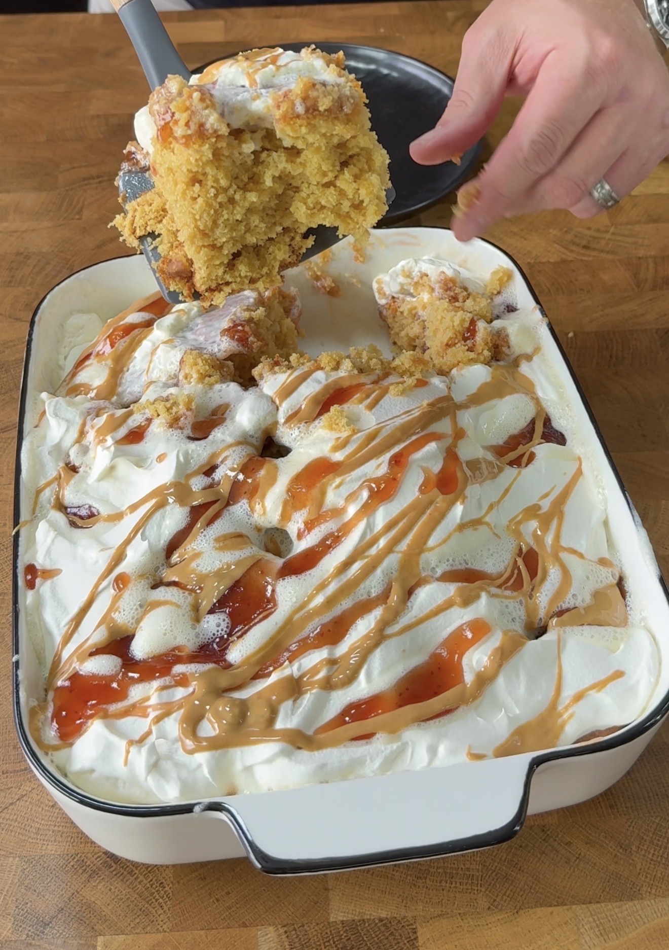 A hand lifts a piece of cake from a dish filled with fluffy whipped topping, caramel and strawberry sauces drizzled over the surface. The cake appears moist and golden, set on a wooden countertop.