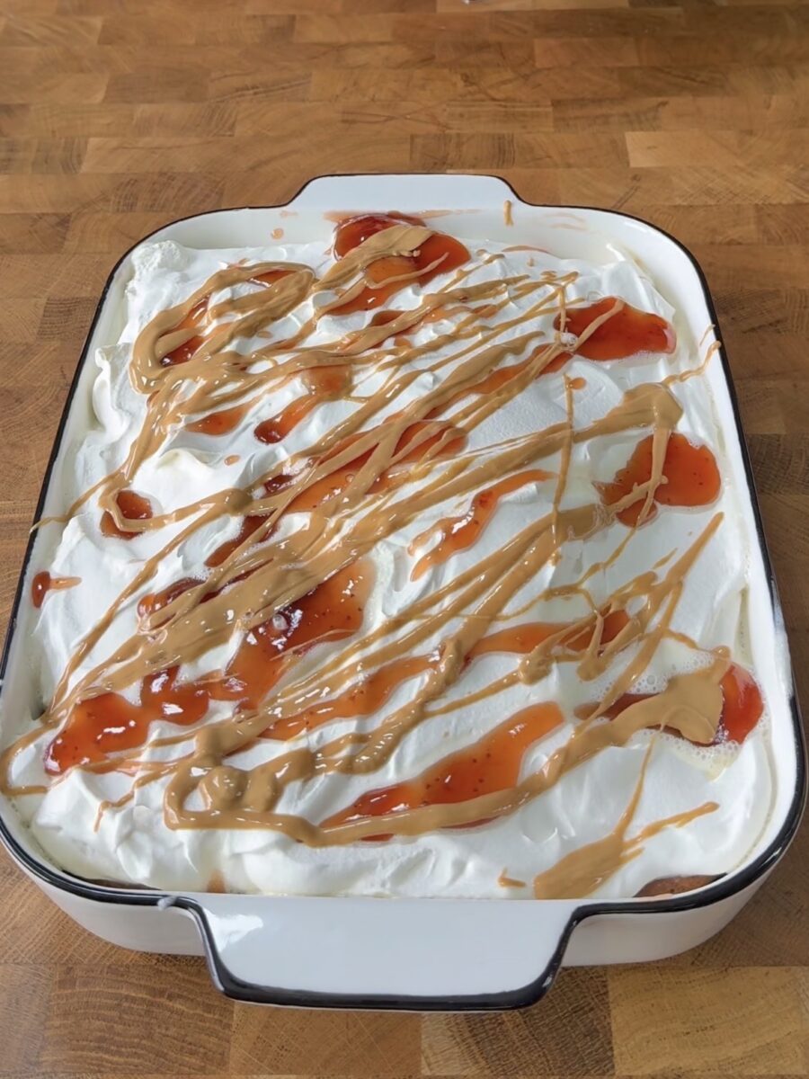 A rectangular dish filled with a layer of whipped cream, topped with drizzles of peanut butter and swirls of red jelly, sits on a wooden surface.
