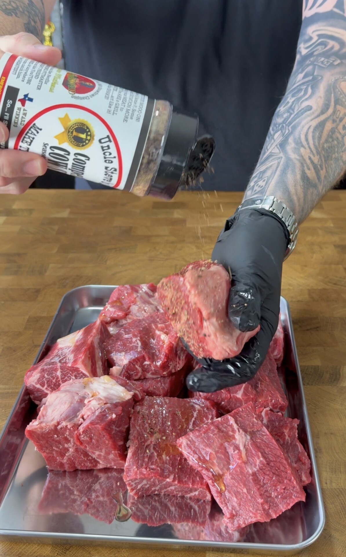 A tattooed person wearing a black glove seasons a tray of raw beef chunks with Uncle Steves Shake, holding one chunk above the tray on a wooden surface.