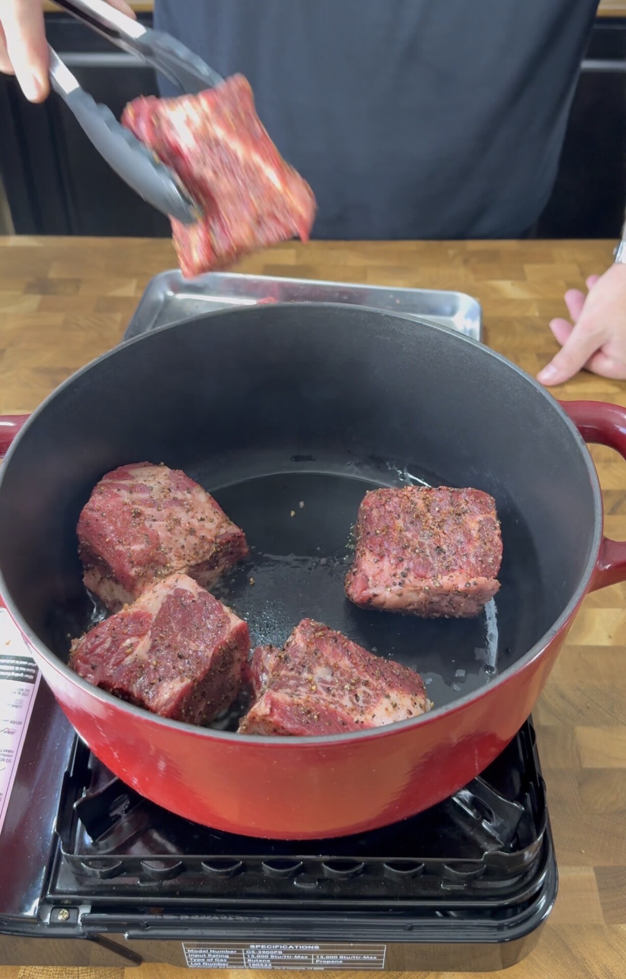 Four seasoned beef short ribs are being seared in a large red Dutch oven on a stovetop. A person is using tongs to add another piece of beef to the pot. The scene is set on a wooden countertop.