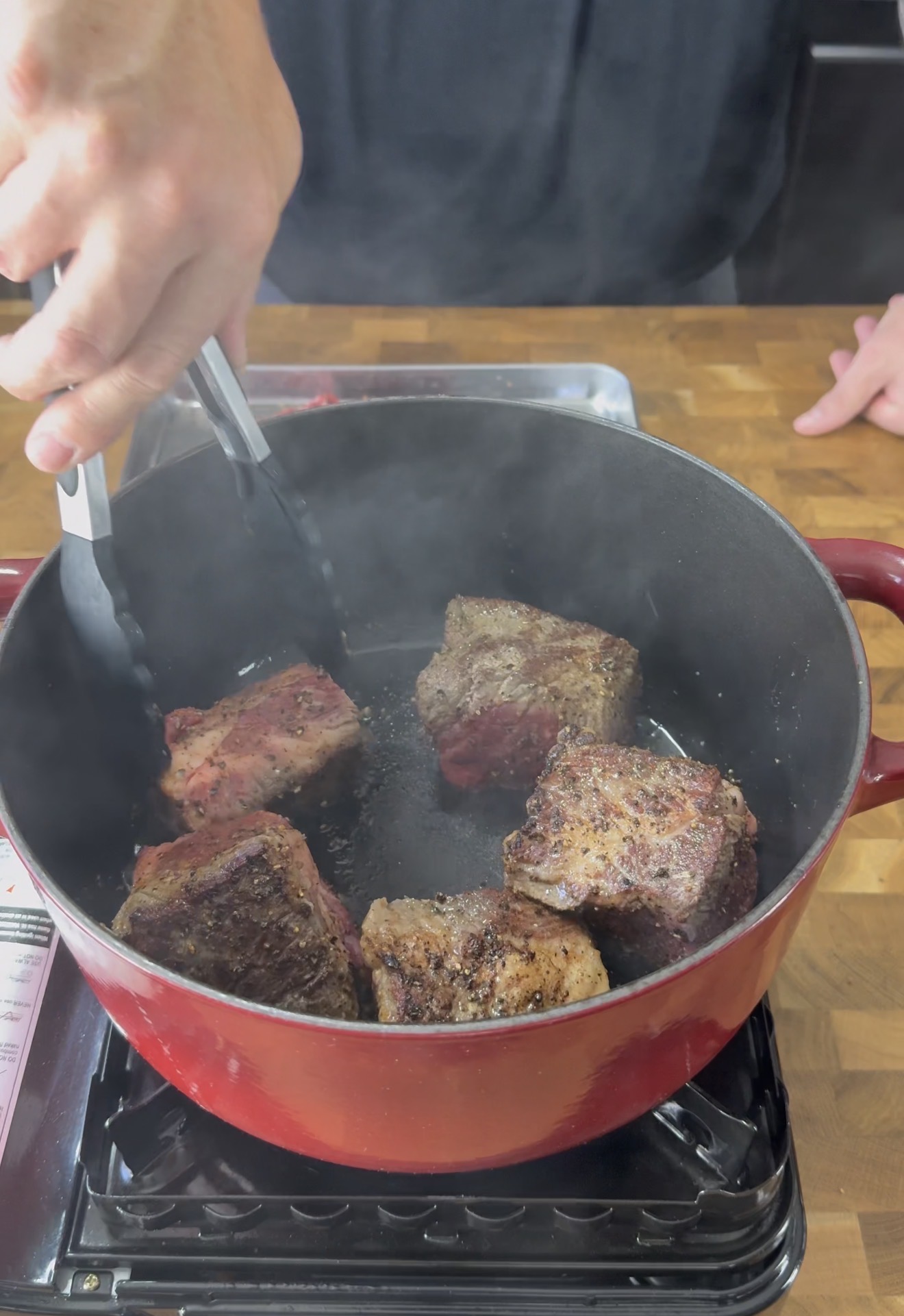 A person uses tongs to sear large chunks of seasoned beef in a red Dutch oven on a stovetop, with steam rising as the meat browns.