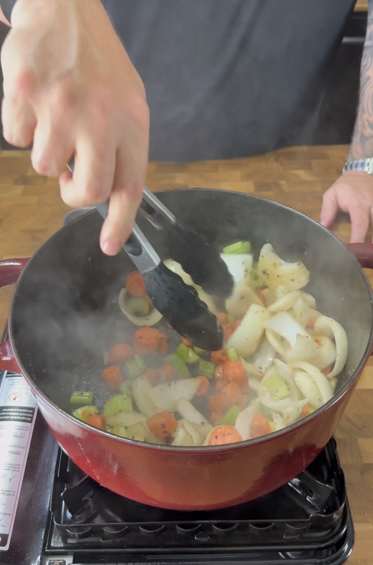 A person uses tongs to stir chopped onions, carrots, and celery in a red pot on a stovetop; steam rises from the cooking vegetables.
