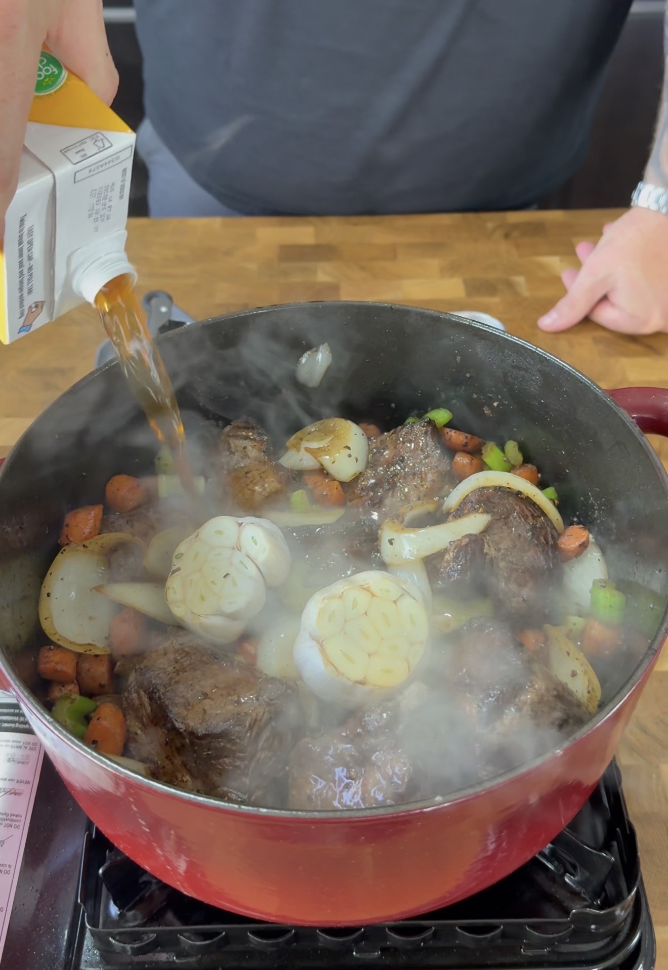 A person pours broth into a red pot filled with browned meat, halved garlic bulbs, onions, carrots, and celery, as steam rises from the cooking ingredients on a stovetop.