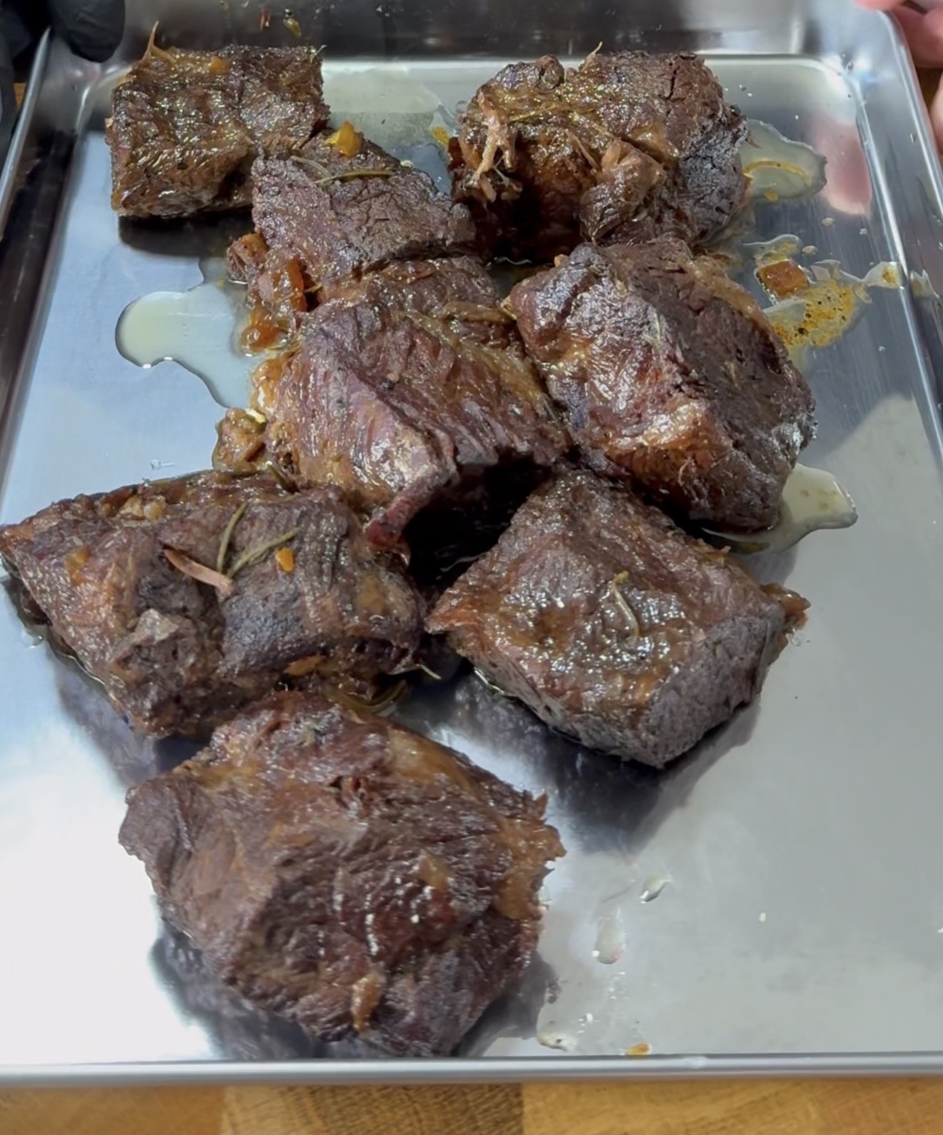 Chunks of cooked, browned beef resting on a shiny metal tray, glistening with juices and bits of seasoning.