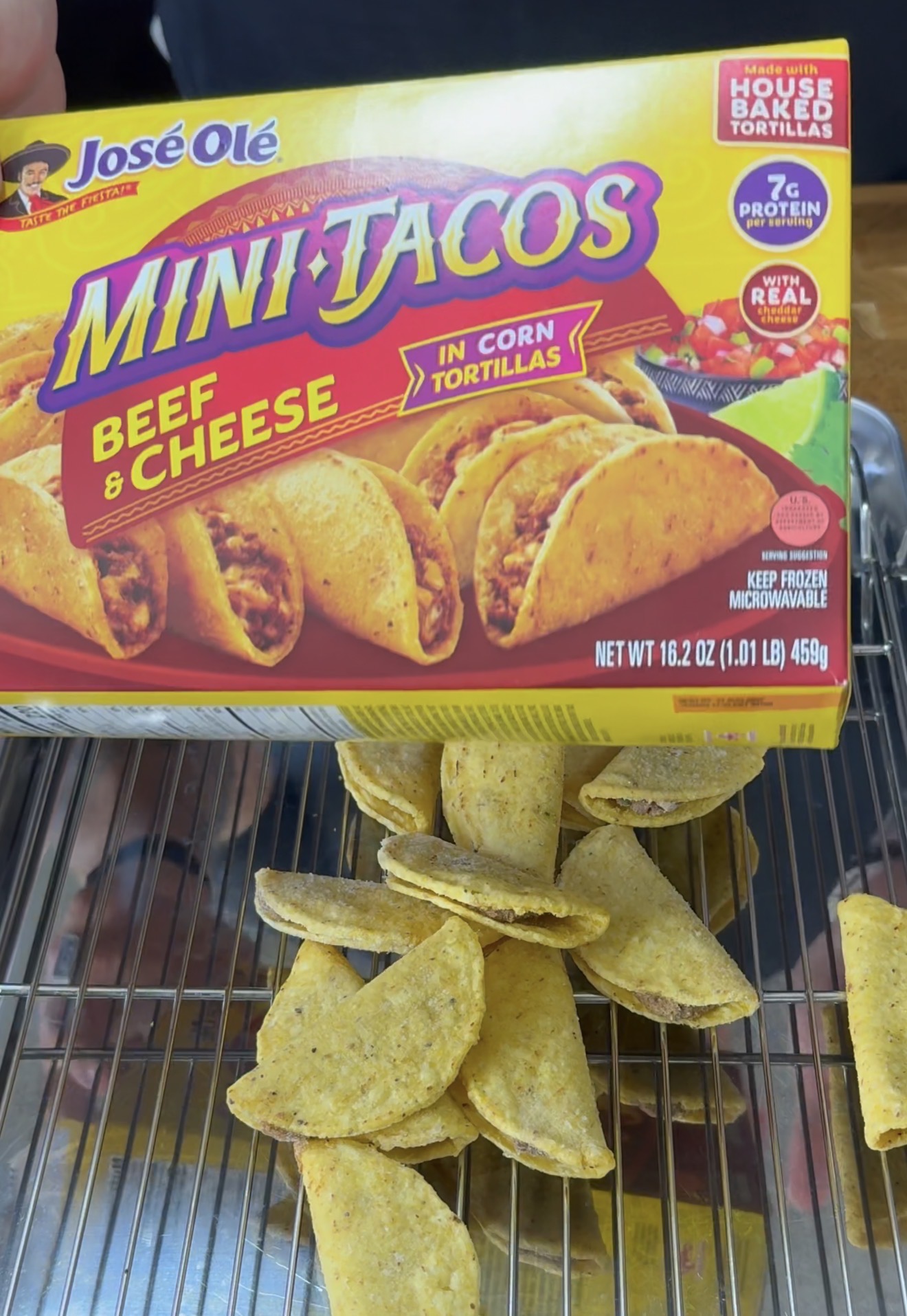 A hand holds a José Olé Mini Tacos Beef & Cheese box above a tray of frozen mini tacos arranged on a metal rack.