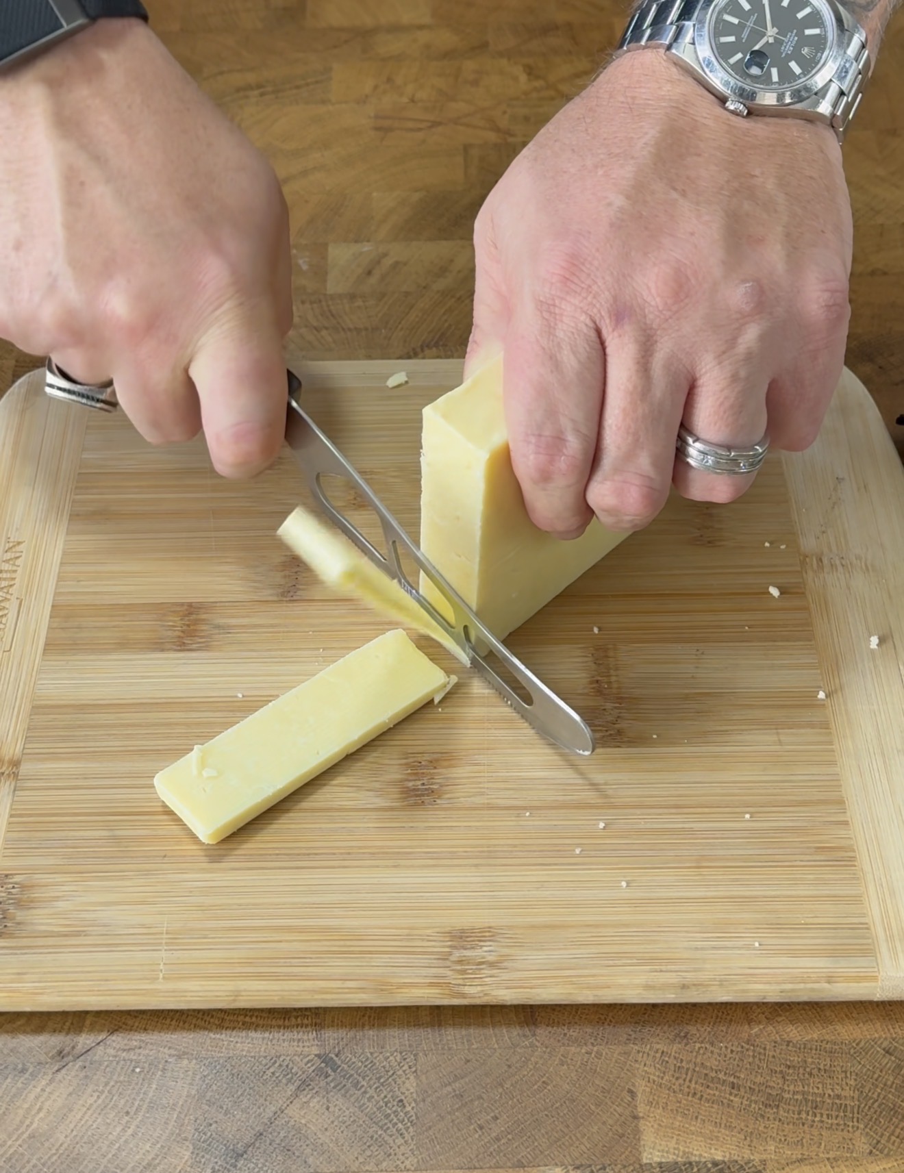 A person’s hands slicing a block of cheese on a wooden cutting board with a knife. The person is wearing a watch and a ring.