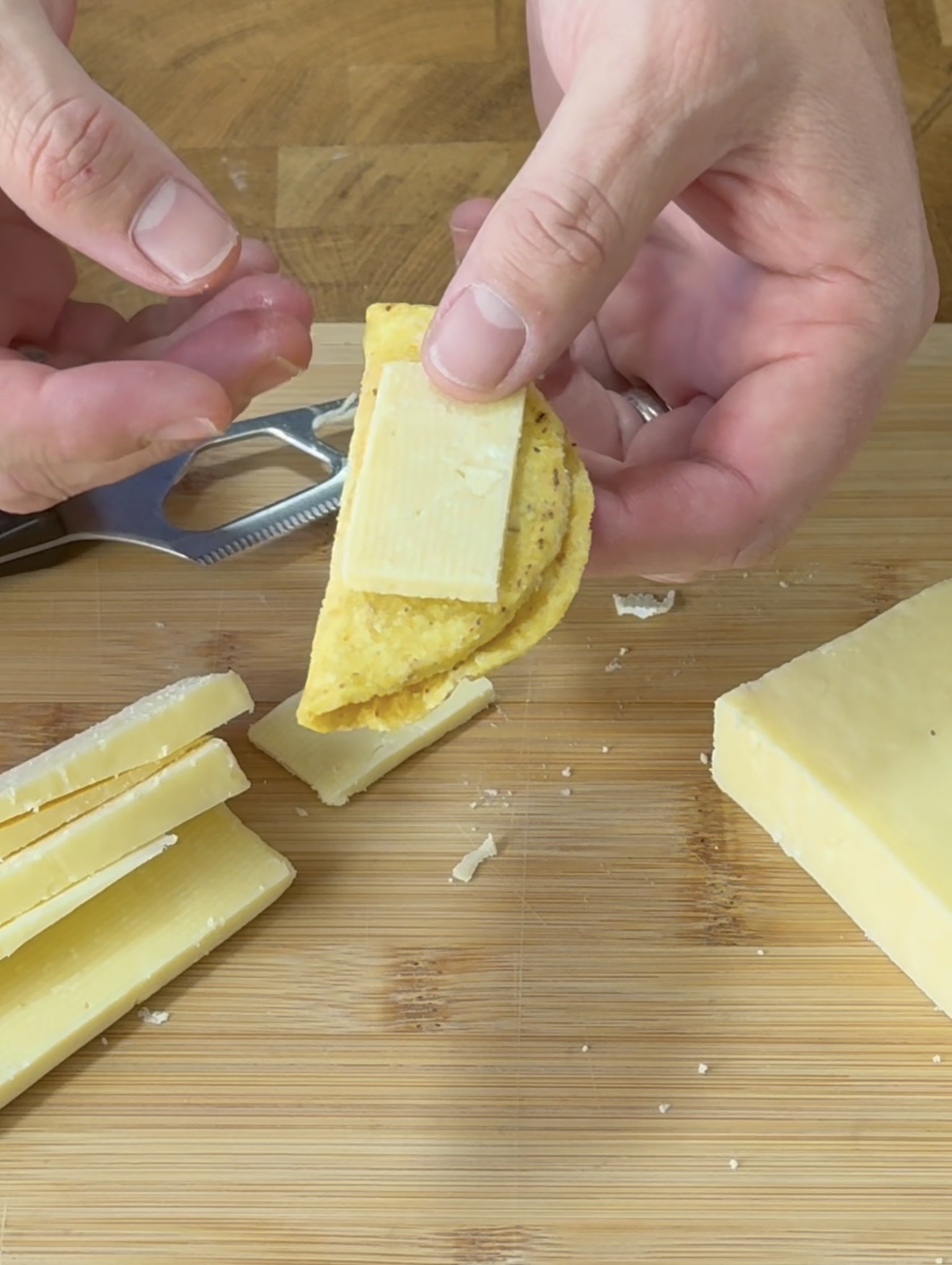 A person folds a corn tortilla around a slice of cheese over a wooden cutting board. Several cheese slices and a block of cheese are nearby.