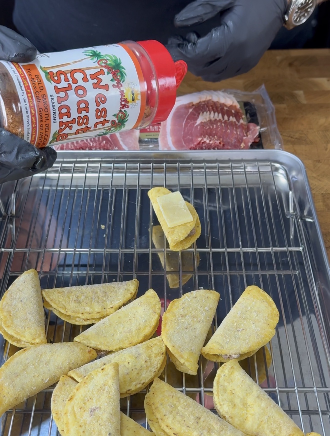 A person wearing black gloves sprinkles West Coast Shake seasoning over folded taco shells on a wire rack, with sliced bacon and cheese also visible on the counter.