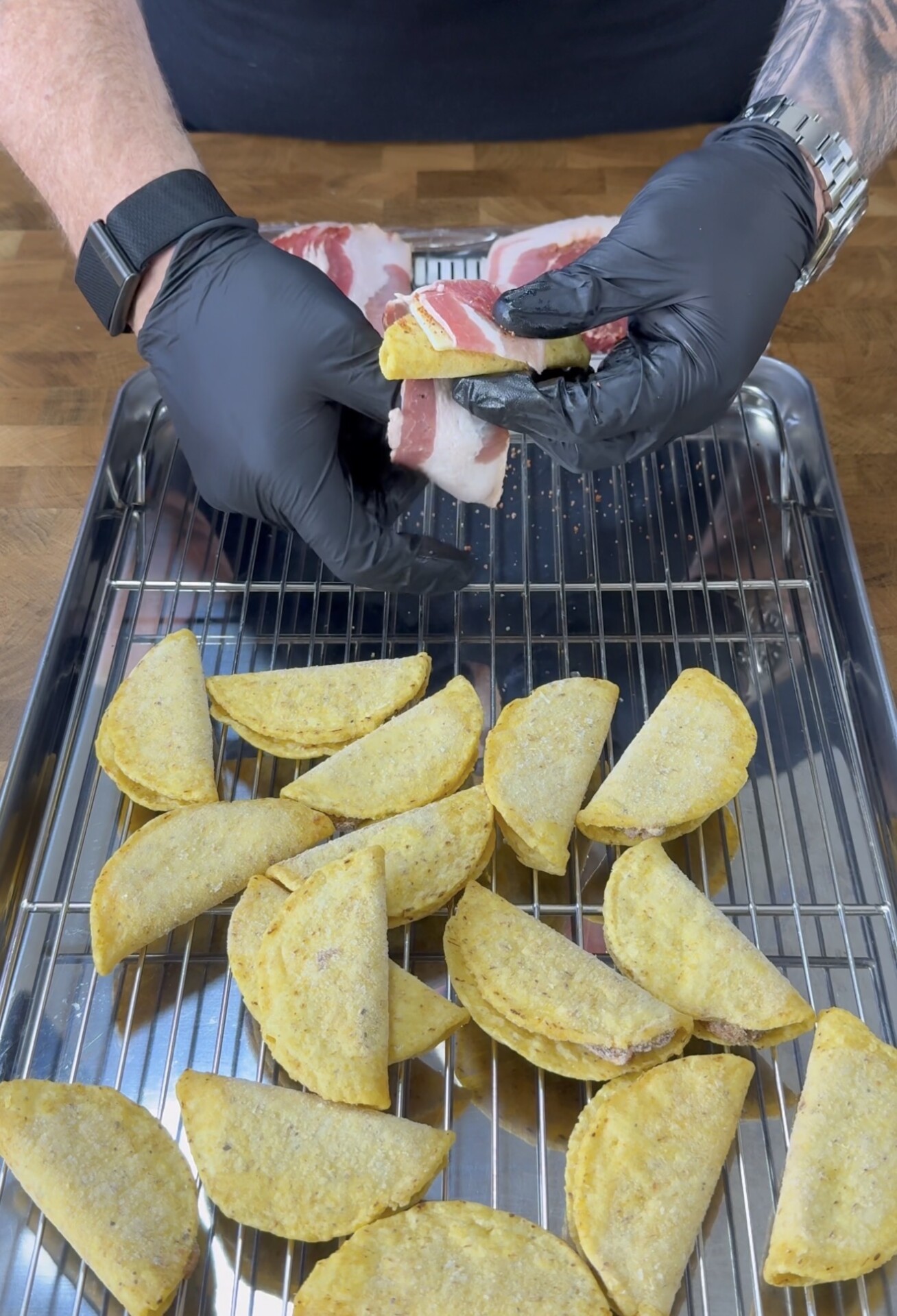 A person wearing black gloves wraps bacon around mini taco shells on a wire rack over a metal tray, with several mini tacos already arranged on the rack.