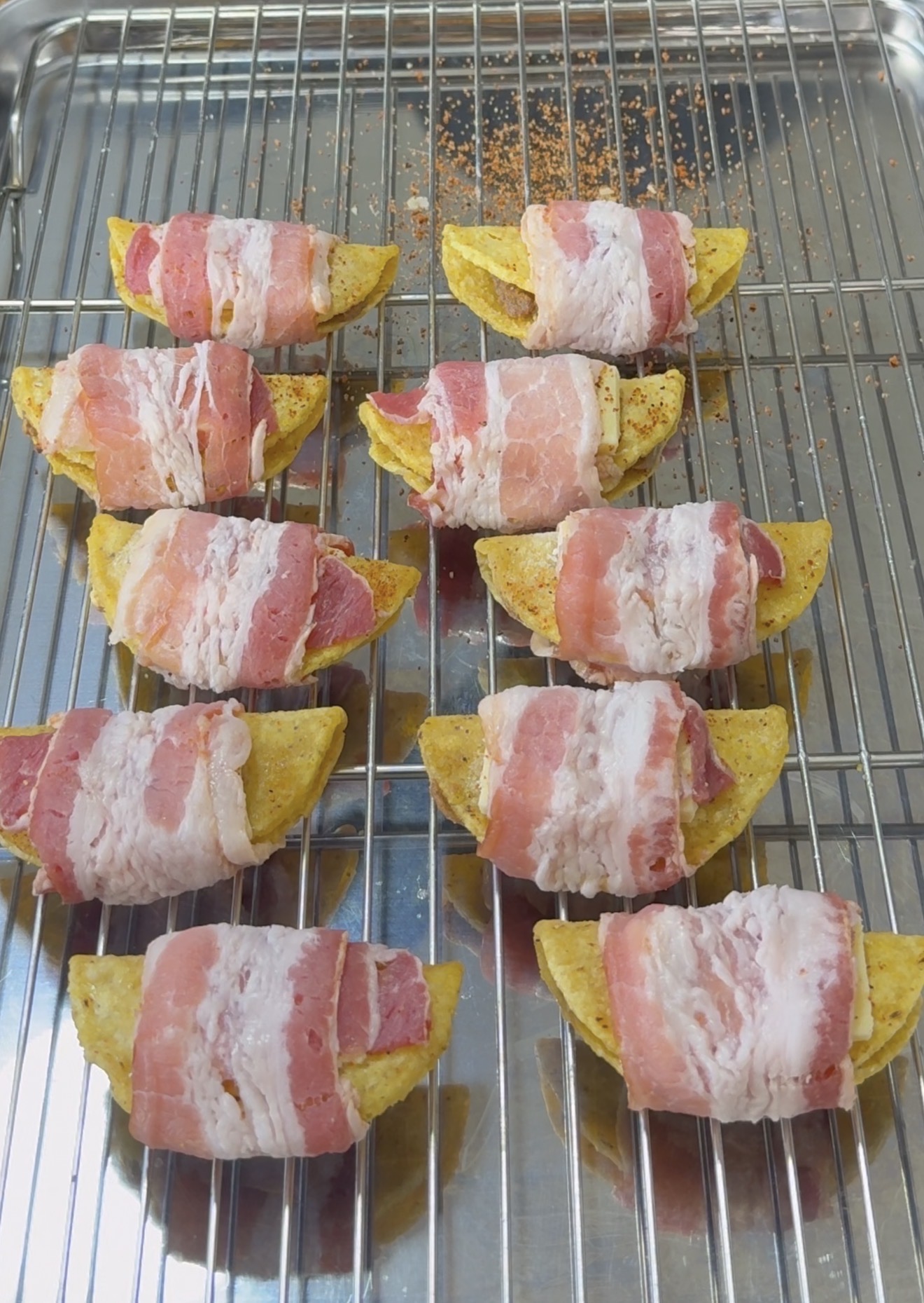 Ten potato wedges wrapped in raw bacon are arranged on a metal cooling rack placed over a baking tray, ready to be cooked.