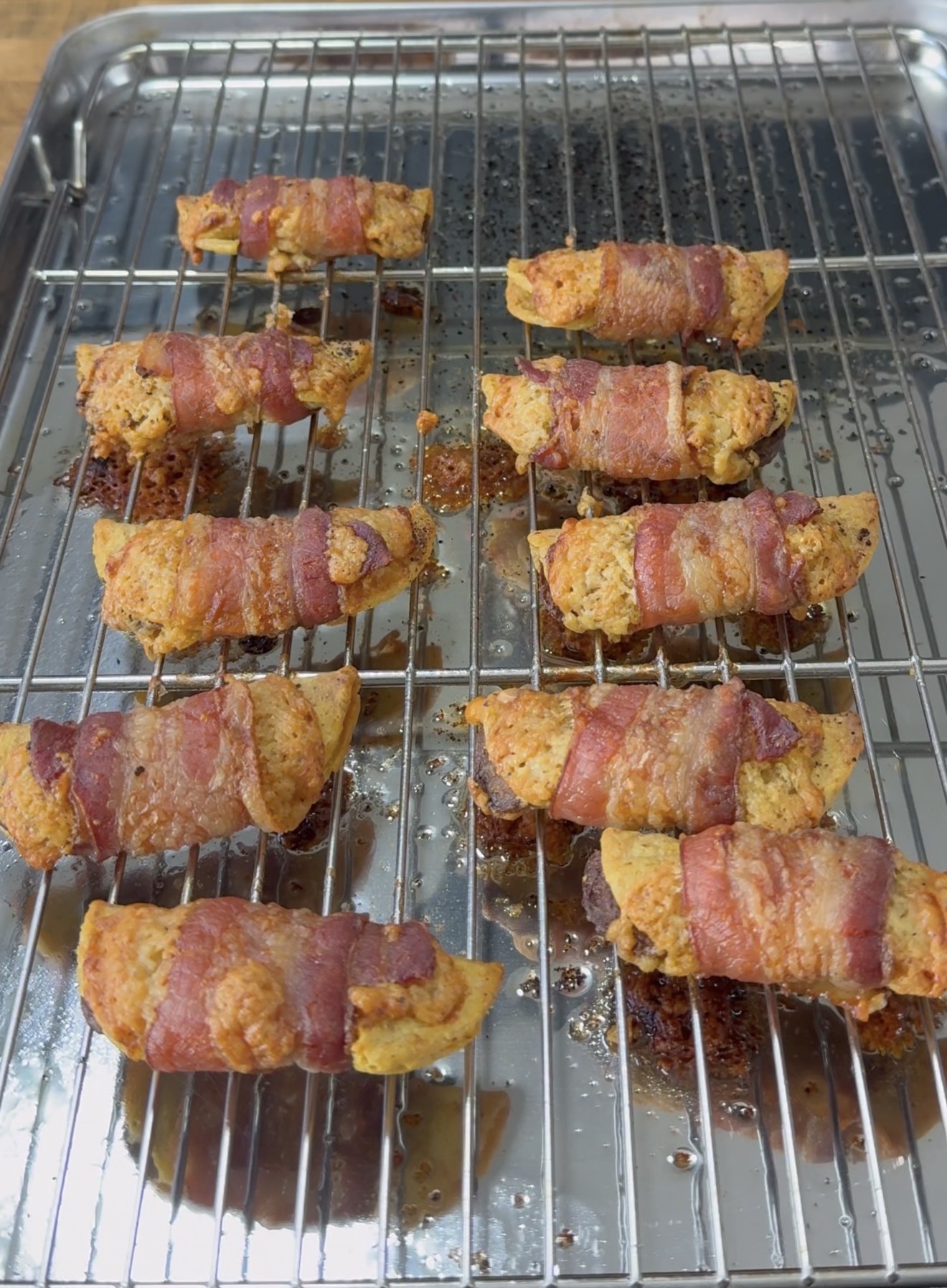 Ten pieces of chicken wrapped in bacon are arranged on a wire rack over a baking sheet, freshly cooked and lightly browned with some drippings on the tray below.