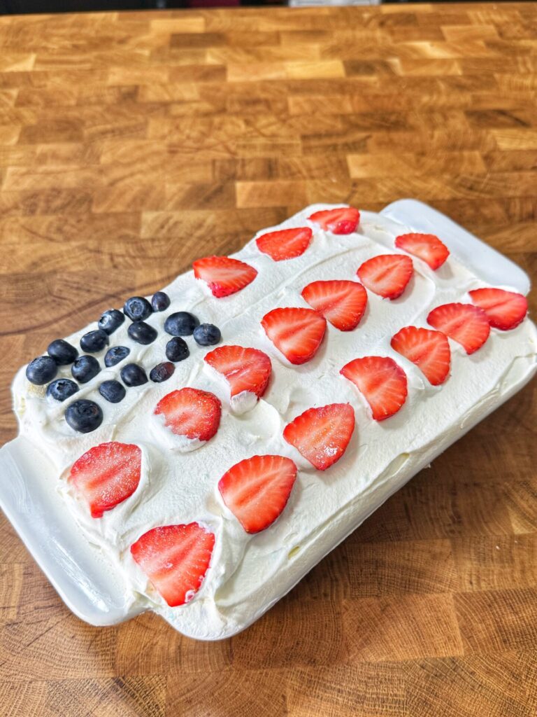 A rectangular cake with white frosting, decorated with blueberries in the corner and rows of sliced strawberries, resembling an American flag, sits on a wooden surface.