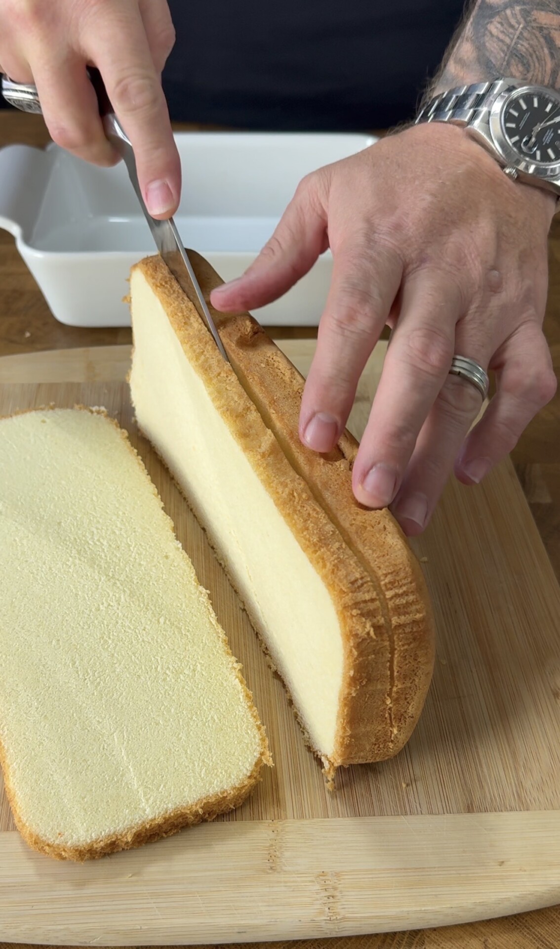 A person slices a loaf of pound cake on a wooden cutting board, with one large slice already cut. Their hands are visible, one holding the cake and the other guiding the knife. A baking dish is in the background.
