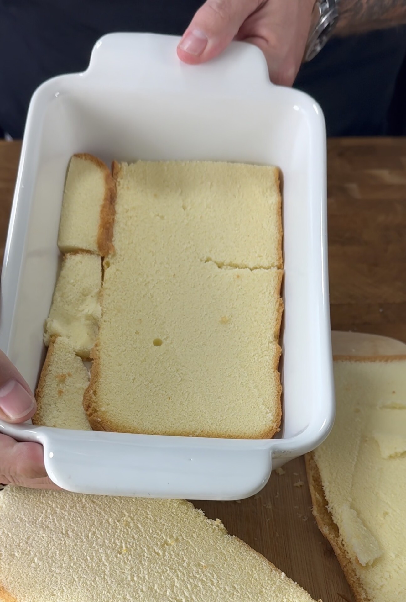 A person places slices of plain sponge cake into a white rectangular baking dish, pressing them to cover the bottom evenly.