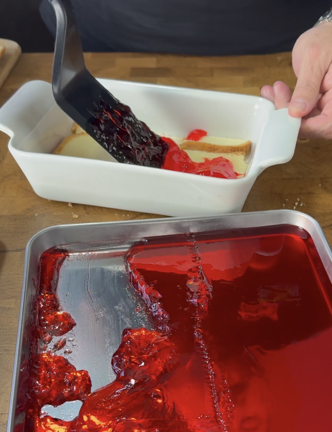 A person uses a spatula to scoop red gelatin from a metal tray and layer it onto a cake or dessert in a white ceramic dish on a wooden countertop.