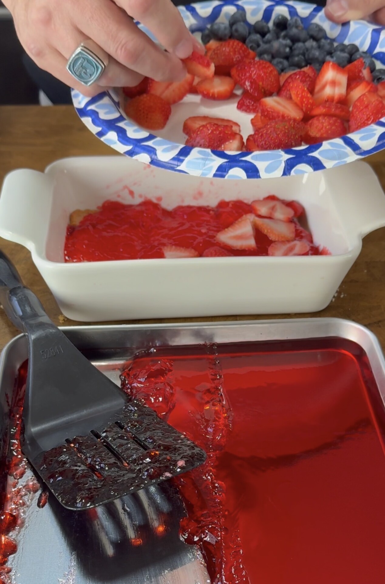 A person adds sliced strawberries and blueberries from a plate into a white baking dish filled with red gelatin. A black spatula and a tray with red gelatin are also visible on the table.
