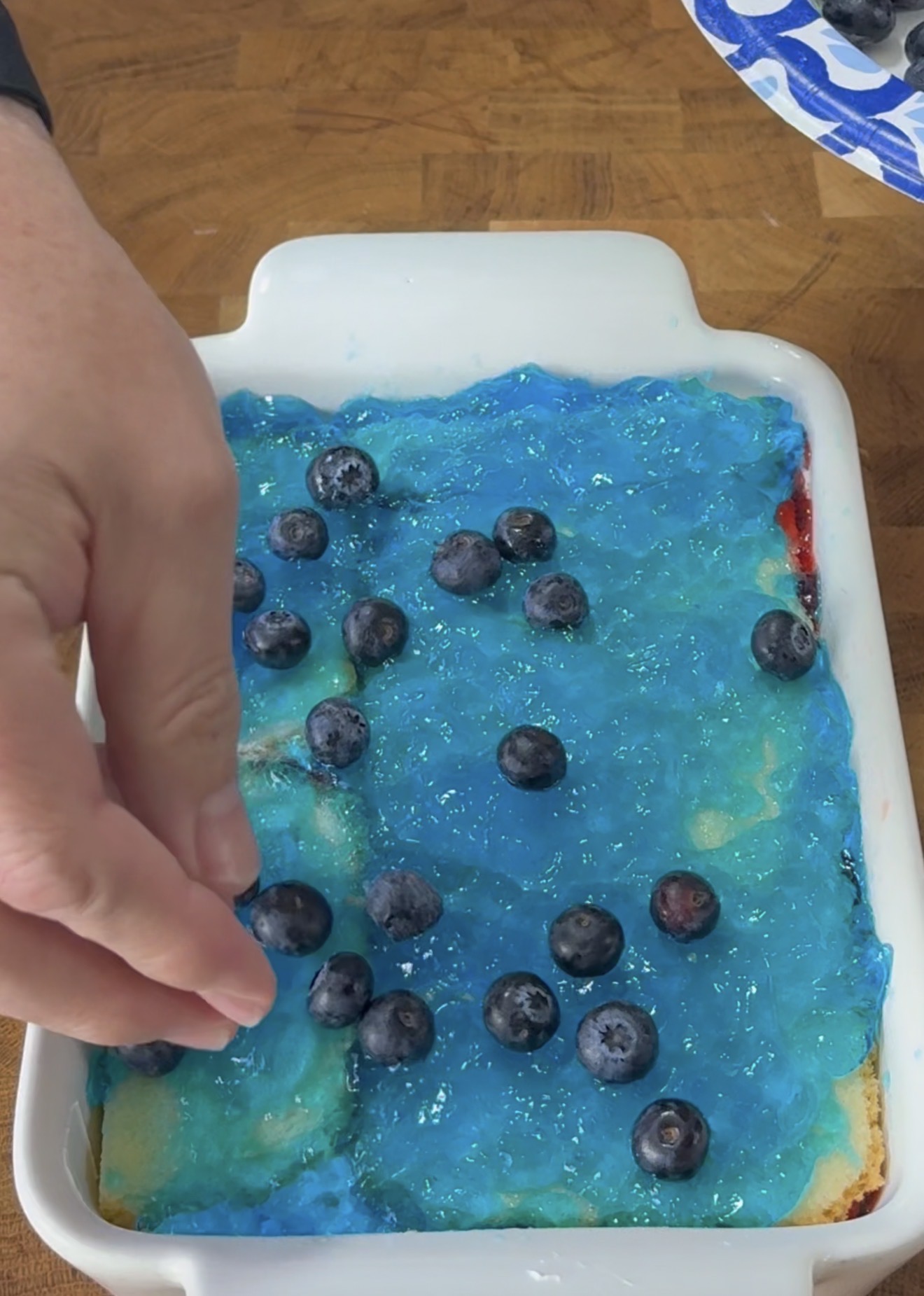 A hand is placing fresh blueberries onto a dessert topped with bright blue gelatin in a white baking dish on a wooden table.