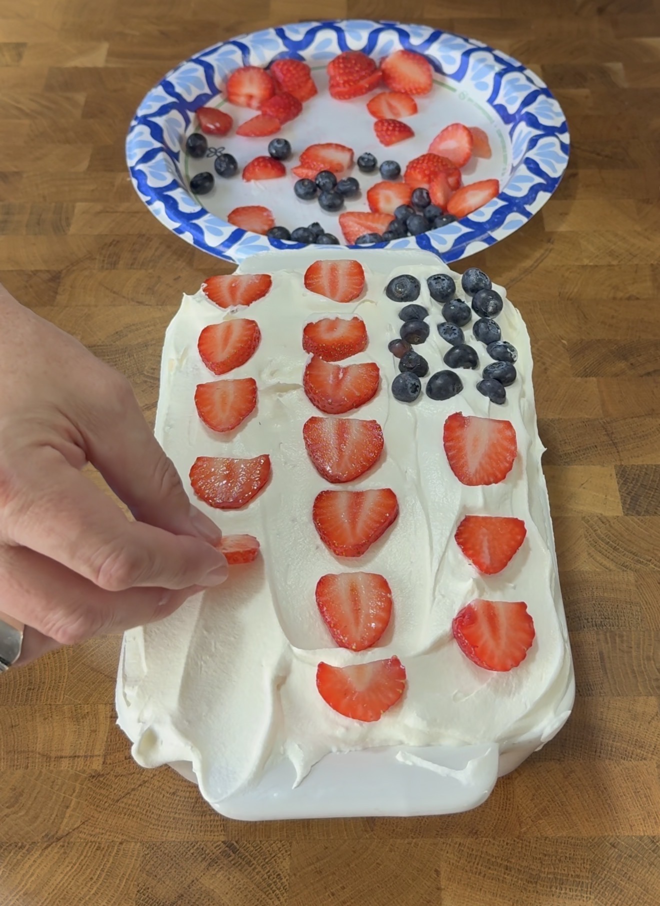 A hand places sliced strawberries on a cake with white frosting. The cake is decorated to resemble an American flag with strawberry stripes and a corner of blueberries. A paper plate with extra berries is in the background.