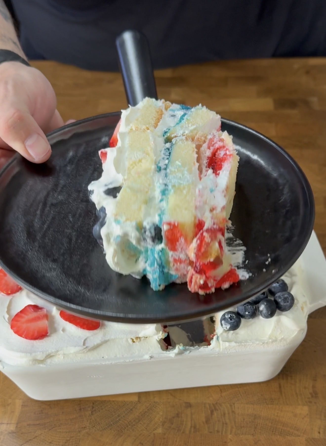A person serves a slice of layered cake with red, white, and blue colors and whipped cream onto a black plate. The cake is topped with strawberries and blueberries on a wooden surface.