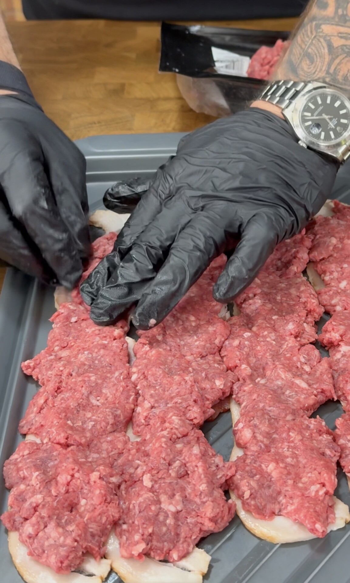 A person wearing black gloves spreads raw ground beef over sliced bread on a baking tray. The person has a tattooed arm and is wearing a watch.