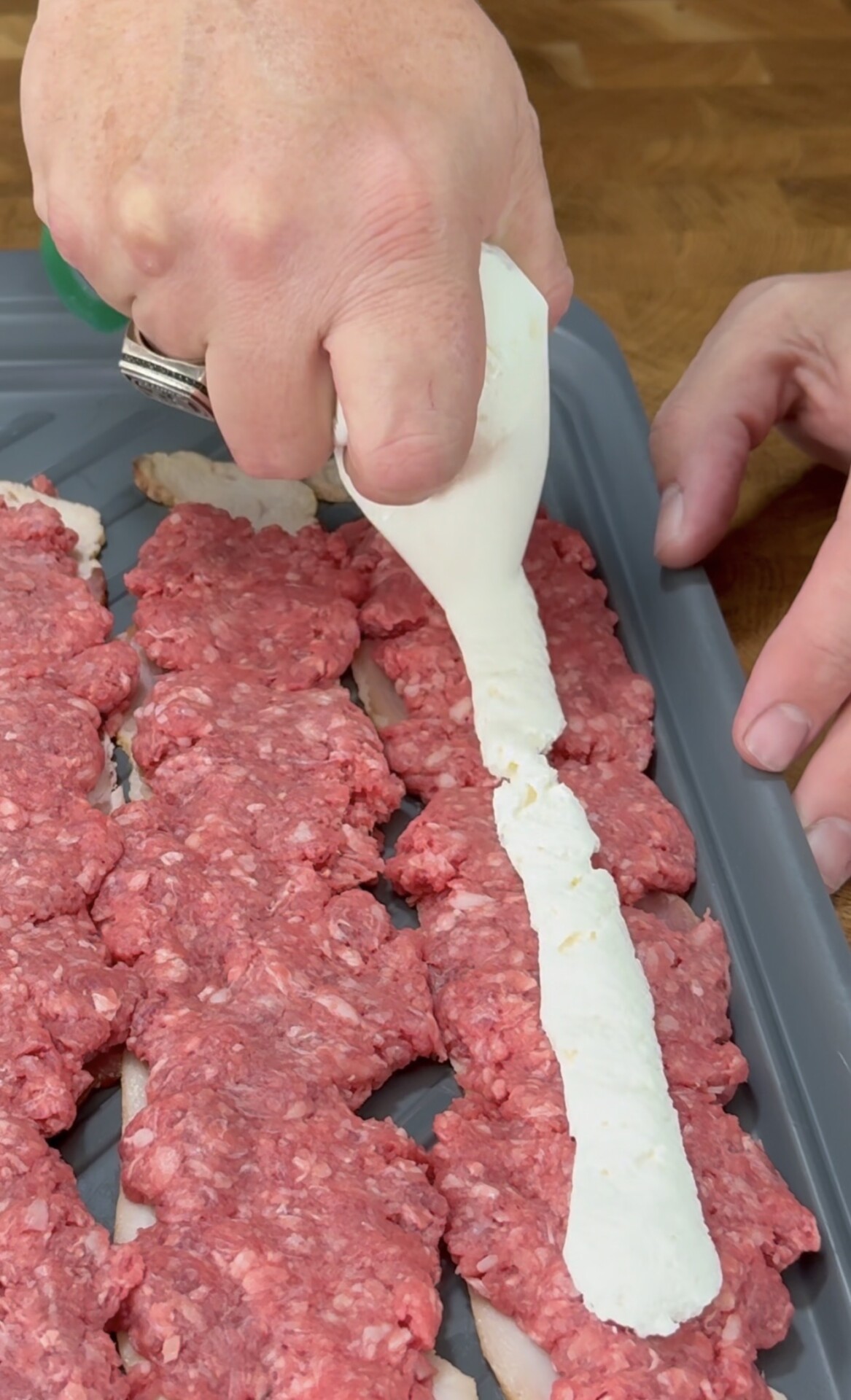 A hand pipes a white creamy mixture onto rows of raw ground beef placed on strips of bacon on a gray tray.